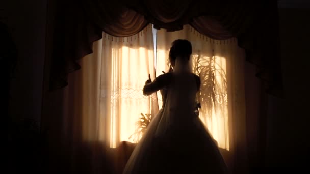 Silhouette of the bride near the window. Slowly opens the curtains and the rays of the sun fall into the room — Stock Video