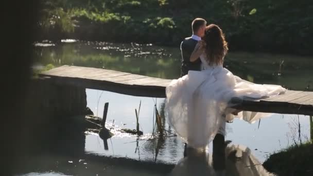 A menyasszony ülni egy fából készült hídon a folyó közelében. Boldog családi pár, kiadási időt a parkban — Stock videók