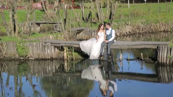 Las novias se sientan en un puente de madera cerca del río. Pareja feliz familia, pasar tiempo en el parque — Vídeos de Stock