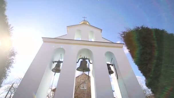 Церковные колокола в солнечный день при сильном ветре. Ветер трясет деревья возле колокольни, а Сонечка светит в келье возле церковных колоколов. — стоковое видео