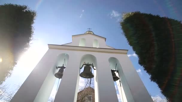 Церковные колокола в солнечный день при сильном ветре. Ветер трясет деревья возле колокольни, а Сонечка светит в келье возле церковных колоколов. — стоковое видео
