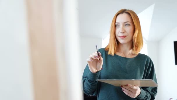 Vrouw kunstenaar schetst een beeld. De kunstenaar heeft een doordachte expressie. Het proces van het schilderen van een foto, een vrouw op zoek naar inspiratie om te tekenen — Stockvideo