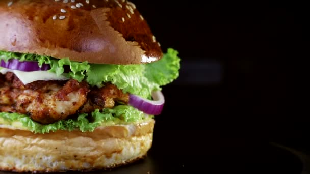 Fast food, close-up. Hambúrguer com costeletas assadas, verdes, tomate e cebola, girando lentamente em uma tábua de madeira em um fundo preto — Vídeo de Stock