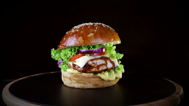 Comida rápida. Hambúrguer com costeletas assadas, verdes, tomate e cebola, girando lentamente em uma tábua de madeira em um fundo preto — Vídeo de Stock