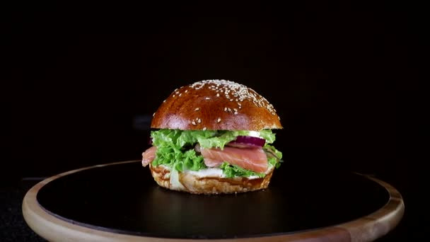 Comida rápida. Hamburguesa con pescado rojo, salmón y trucha, tomate y cebolla, girando lentamente sobre un tablero de madera sobre un fondo negro — Vídeo de stock