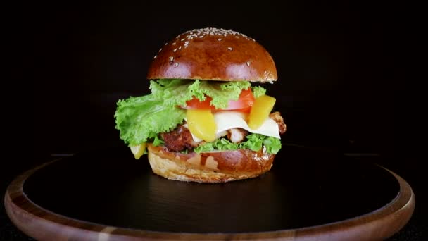 Hambúrguer com um grande pedaço de carne, verdes e queijo estão girando em uma placa de madeira em fundo preto — Vídeo de Stock