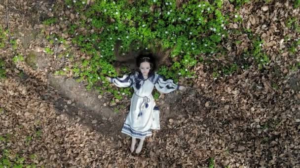 Vista aérea en la cima. Una niña está acostada en la hierba con flores blancas con los ojos cerrados. La cámara se aleja lentamente y se acerca. Disparo de un dron — Vídeos de Stock