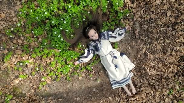 Вид сверху с воздуха. Маленькая девочка лежит на траве с белыми цветами с закрытыми глазами. Камера медленно отходит и приближается. Выстрел из беспилотника — стоковое видео