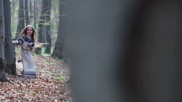 Pequena menina descalça corre em uma floresta ou parque e se esconde atrás de árvores em um vestido branco com padrões azuis . — Vídeo de Stock