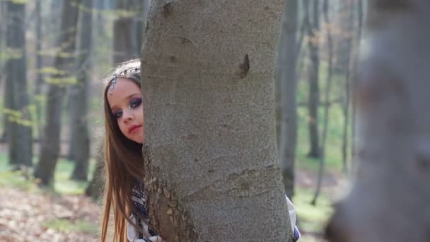 Piccola ragazza a piedi nudi corre in una foresta o parco e si nasconde dietro gli alberi in un abito bianco con motivi blu . — Video Stock