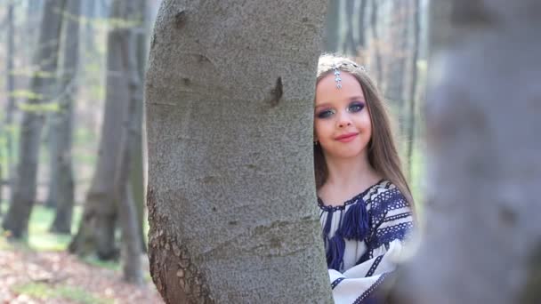 Kleine Barefoot meisje loopt in een bos of Park en verbergt achter bomen in een witte jurk met blauwe patronen. — Stockvideo