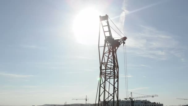 Dva stavební jeřáby na pozadí modrého nebe a slunce, které svítí v kameře za stavebním jeřábem — Stock video
