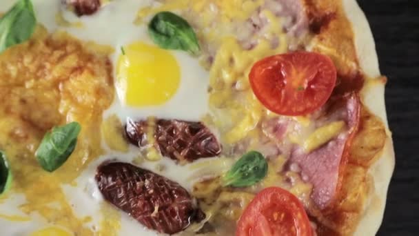 Close-up van een pizza met kaas tomaten, worst en ei die draait om zichzelf op een zwarte achtergrond uitzicht vanaf de top — Stockvideo