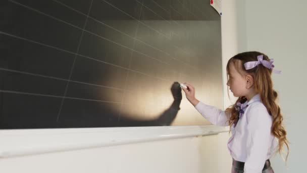 Kislány egy iskolai osztályteremben a tábla közelében rajzol az iskola táblájára fehér krétával. — Stock videók