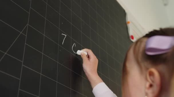 Una niña pequeña en un aula de la escuela en la junta resuelve un ejemplo. Alumno con tiza blanca escribe un ejemplo matemático en el primer plano del tablero . — Vídeos de Stock