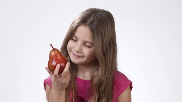 Una niña sobre un fondo blanco sostiene en su mano una pera fragante madura. La chica muerde y come una deliciosa pera roja . — Vídeos de Stock