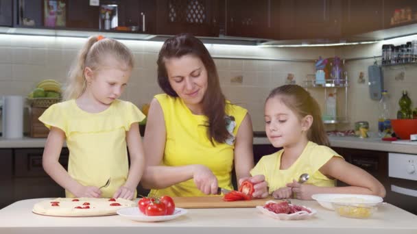 Mom and daughters slice a tomato in the kitchen to make pizza. Children cook pizza with tomatoes and other ingredients together with their mother. — Stock Video