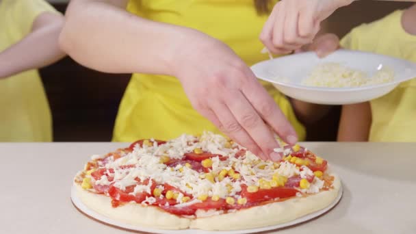 Copiii împreună cu mama gătesc pizza aproape. Fetele stropesc pizza cu brânză de aproape și o ajută pe mama să gătească în bucătărie . — Videoclip de stoc