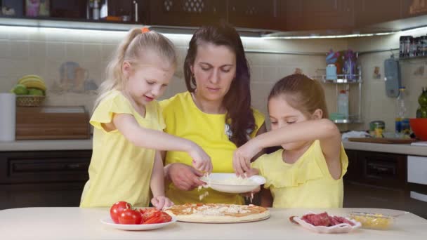 Barnen lagar pizza med sin mamma. Flickorna strö pizza med ost och hjälpa sin mamma laga mat i köket. — Stockvideo