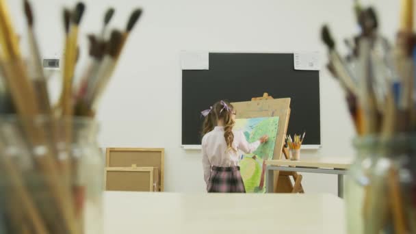 La niña pinta un cuadro y mira a la cámara. Niña feliz sonriendo en el fondo de sus pinturas de dibujo . — Vídeos de Stock