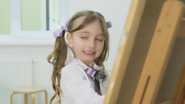 Menina pinta um quadro e olha para a câmera. Menina feliz sorrindo no fundo de suas pinturas de desenho . — Vídeo de Stock