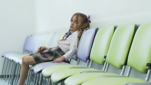 Triste bambina seduta da sola su una sedia e triste. Depressione nei bambini piccoli a scuola. La ragazza è chiusa in se stessa seduta nel corridoio su sedie con le gambe pressate insieme . — Video Stock