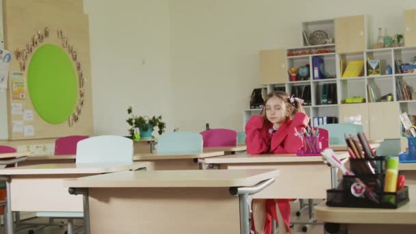 Een kind op school zit aan een bureau, doet zijn haar recht en kijkt naar de camera. — Stockvideo