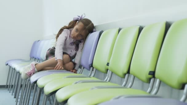 Triest klein meisje zit alleen in een stoel en verdrietig. Depressie bij jonge kinderen op school. Het meisje zit in zichzelf opgesloten in de gang op stoelen met haar benen tegen elkaar gedrukt. — Stockvideo