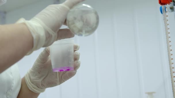 Een laboratoriummedewerker giet een roze substantie uit een glazen kolf en schudt deze in een plastic maatbeker. De onderzoeker voert een experiment uit in het laboratorium — Stockvideo