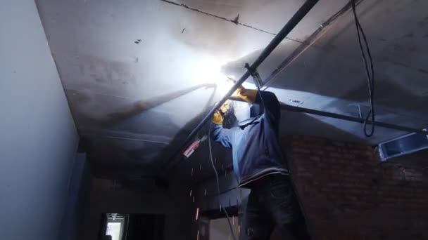 Um soldador em um canteiro de obras solda um tubo de ferro sob o teto da sala. O soldador trabalha em um quarto escuro em um canteiro de obras . — Vídeo de Stock