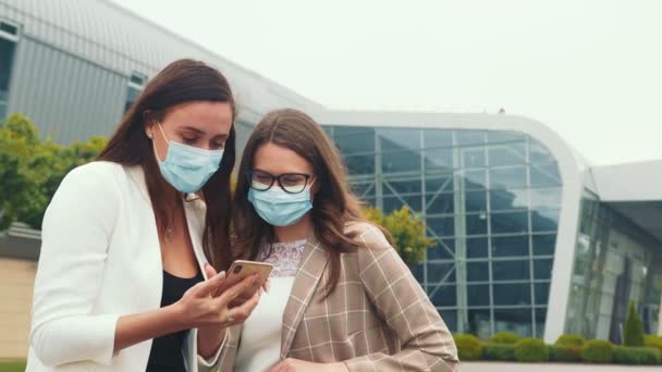 Dos chicas con máscaras médicas con un teléfono móvil navegar por el contenido del teléfono y reír emocionalmente. Emociones de las niñas mientras mira un teléfono móvil . — Vídeos de Stock