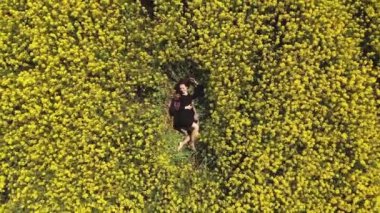 Siyah elbiseli genç kızın hava manzarası kolza ve sarı çiçeklerle dolu bir tarlada yatıyor. Peri tarlasında gözleri kapalı yatan bir kadın. Kamera kızdan uzaklaşıyor..