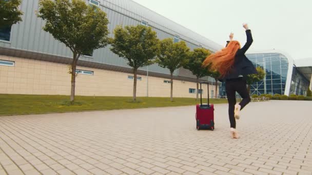 Щаслива молода жінка танцює біля багажної валізи на фоні терміналу аеропорту. Жінка з рудим волоссям жаліє і показує свої емоції, стрибаючи і бігаючи навколо свого багажу . — стокове відео
