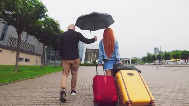 Жінка перевозить дві валізи до терміналу аеропорту і чоловік, який тримає парасольку. Пріоритет чоловіка в сім'ї. Проблеми взаємозв'язку молодої пари . — стокове відео