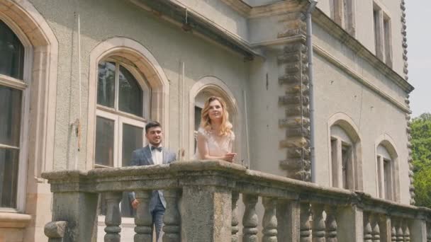 Le marié s'approche de la mariée debout près du balcon en pierre et l'embrasse par derrière. Émotions heureuses d'un jeune couple par une journée ensoleillée. — Video