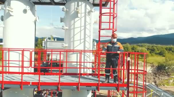 航空视图加油站操作员爬到车站顶部。山区的现代天然气综合体。在山区的背景下，一名煤气工人爬上了煤气管道的梯子。 — 图库视频影像