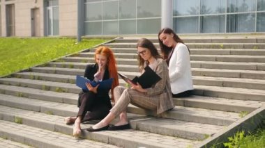 Genç iş kadınları merdivenlerde oturur ve siyah bir klasördeki belgelerin içeriğini inceler ve gülerek eğlenirler. Neşeli kızlar güneşli bir günde gülümsüyor ve iş konuşuyorlar..