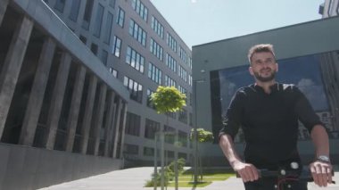 Genç erkek işadamı geleceğin elektrikli scooter 'ıyla modern evler arasındaki parlak güneşte gülümsüyor..