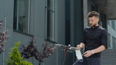 Elektrikli scooter 'ın ekolojik nakliyesinin yanında duran bir adam koruyucu bir tıbbi maske takmak zorunda kalır..