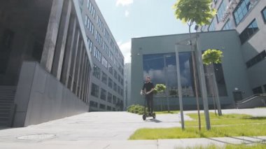 Genç erkek işadamı geleceğin elektrikli scooter 'ıyla modern evler arasındaki parlak güneşte gülümsüyor..