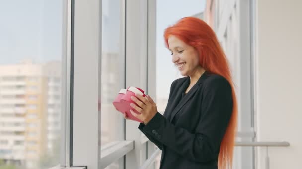 The woman received a gift box in the form of a heart. Happy woman with red hair rejoices at the gift and stands by the window — Stock Video