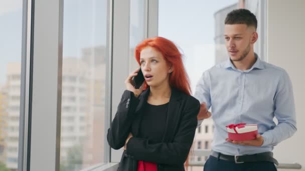 Un uomo fa un regalo a una donna con i capelli rossi. Una donna che parla al telefono riceve un regalo dal marito e si rallegra del regalo ricevuto in ufficio.. — Video Stock