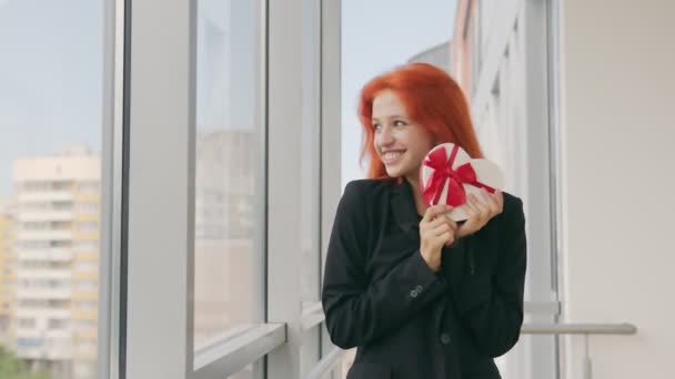 La mujer recibió una caja de regalo en forma de corazón. Mujer feliz con el pelo rojo se regocija en el regalo y se para junto a la ventana — Vídeo de stock
