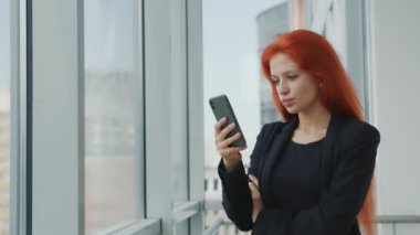 Elinde cep telefonu tutan kızıl saçlı bir kadının portresi. Camdan kasvetli bir şekilde dışarı bakıyor. Ofiste, penceresinin yanında cep telefonu olan kızgın bir kadın var..