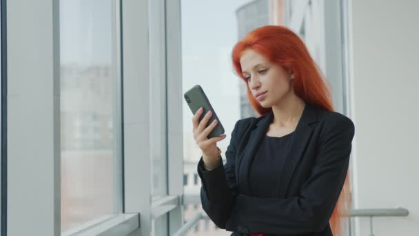 手に携帯電話を持っている赤い髪の女性の肖像画と心から笑顔と窓のそばのオフィスで彼女の髪をまっすぐに — ストック動画