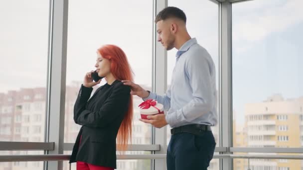 Un uomo fa un regalo a una donna con i capelli rossi. Una donna che parla al telefono riceve un regalo dal marito e si rallegra del regalo ricevuto in ufficio.. — Video Stock