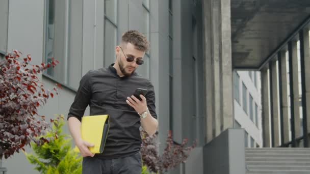 Uomo d'affari con occhiali e una cartella gialla con documenti in attesa di una riunione e guardando il suo orologio. Uomo in attesa di un incontro di lavoro sullo sfondo di edifici moderni nel centro della città. — Video Stock