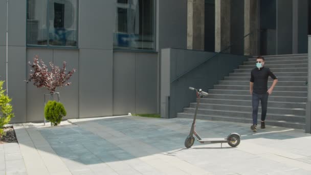 Een jongeman met een medisch beschermend masker die een elektrische scooter gebruikt als modern vervoermiddel in de stad. Een mannelijke zakenman benadert een elektrische scooter en rijdt erop voor zaken. — Stockvideo