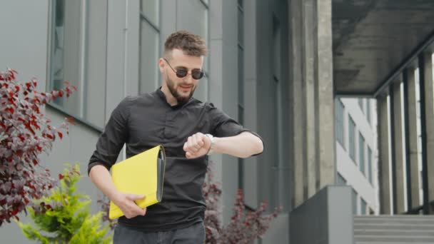 Homme d'affaires avec des lunettes et un dossier jaune avec des documents en attente d'une réunion et en regardant sa montre. Homme en attente d'une réunion d'affaires sur le fond des bâtiments modernes du centre-ville. — Video