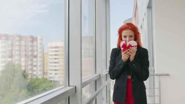 La donna ha ricevuto una scatola di regalo nella forma di un cuore. Donna felice con i capelli rossi gioisce per il regalo e sta vicino alla finestra — Video Stock